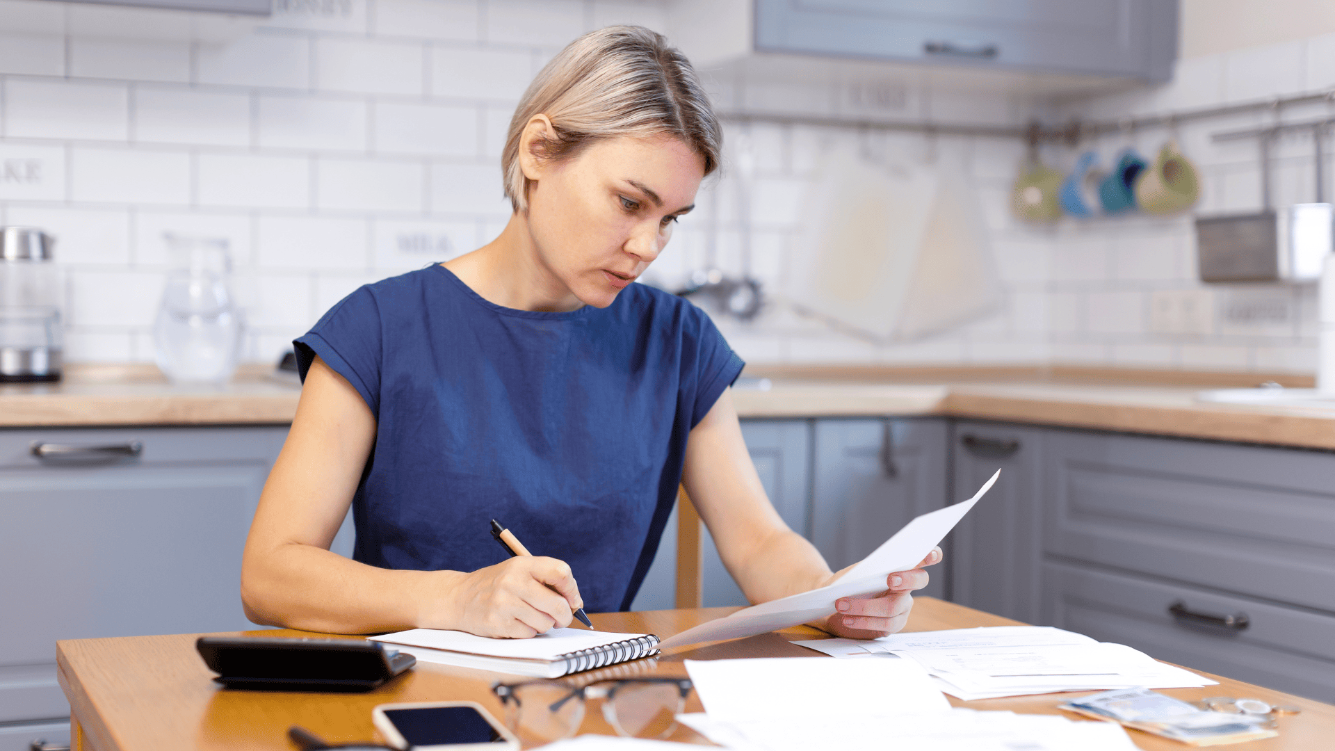 Mujer en la cocina revisando facturas o documentos financieros.