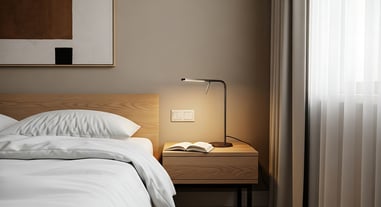 Lámpara encendida sobre mesita de noche con libro abierto, junto a cama con sábanas blancas