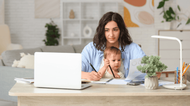 Mujer trabajando desde casa con su bebé en brazos frente a un ordenador portátil