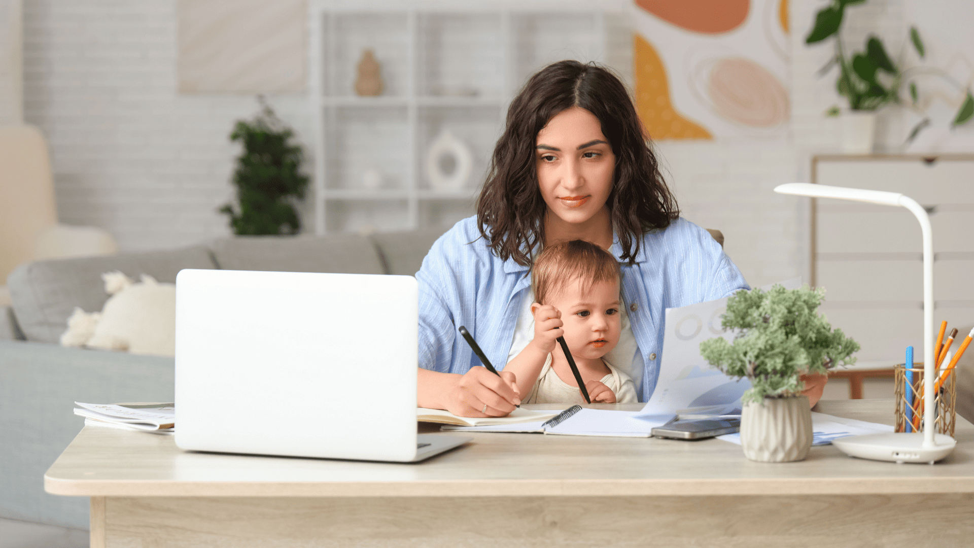Mujer trabajando desde casa con su bebé en brazos frente a un ordenador portátil