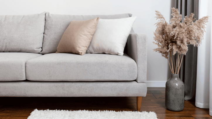 Sofá gris claro con dos cojines y jarrón grande con ramas decorativas junto a una cortina