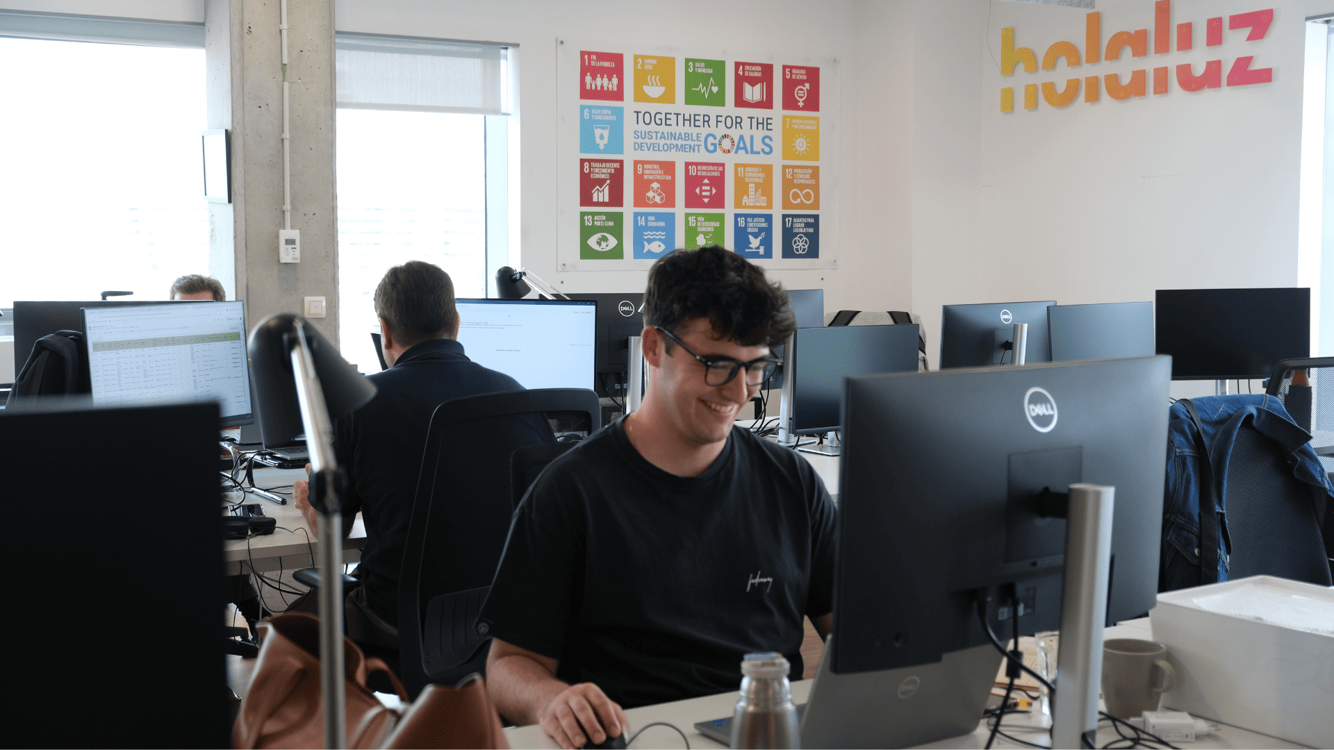 Equipo de Holaluz trabajando en una oficina con varios ordenadores y un cartel de los Objetivos de Desarrollo Sostenible.