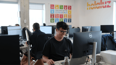 Equipo de Holaluz trabajando en una oficina con varios ordenadores y un cartel de los Objetivos de Desarrollo Sostenible.