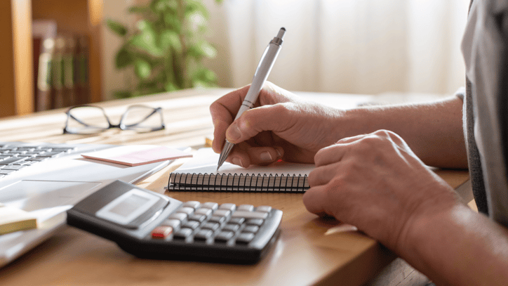 Persona tomando notas en una libreta junto a un ordenador portátil y una calculadora.