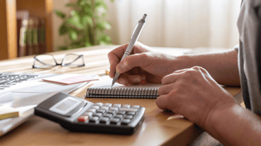 Persona tomando notas en una libreta junto a un ordenador portátil y una calculadora.