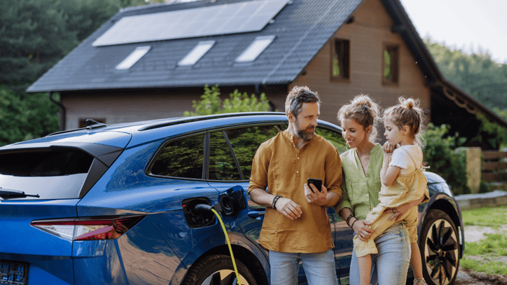 amilia junto a un coche eléctrico en proceso de carga frente a una vivienda con paneles solares