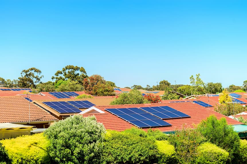 Tejados de casas con tejas rojas equipados con paneles solares, rodeados de vegetación y con un cielo despejado de fondo