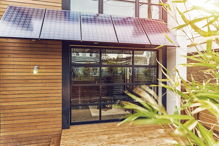 Pared de una casa de madera con una gran ventana y placas solares instaladas en la parte superior como cubierta