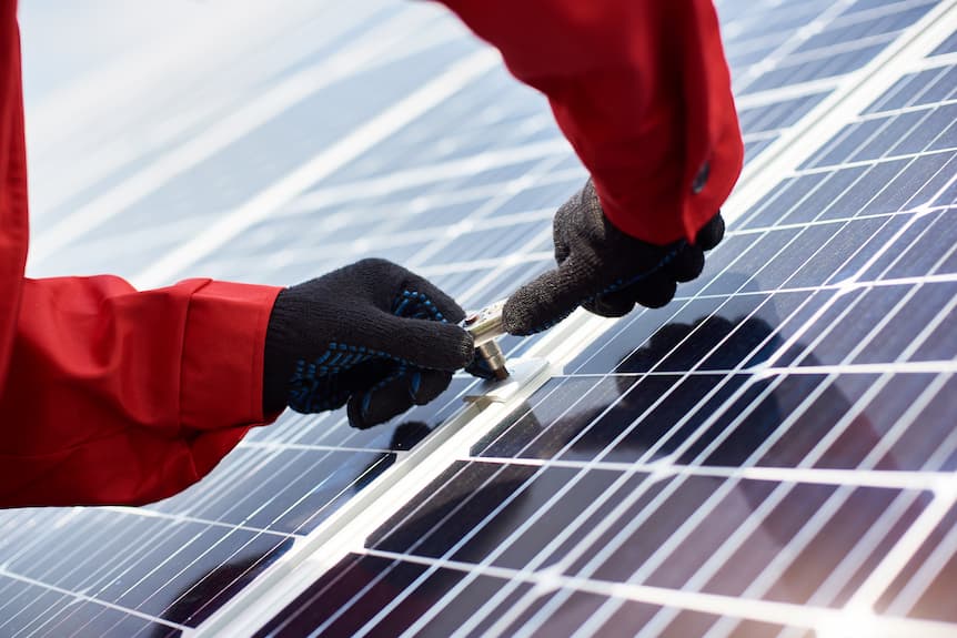 Manos de un trabajador con guantes negros y chaqueta roja ajustando un panel solar con una herramienta