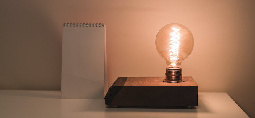 Lámpara de filamento encendida sobre una base de madera junto a un cuaderno en blanco