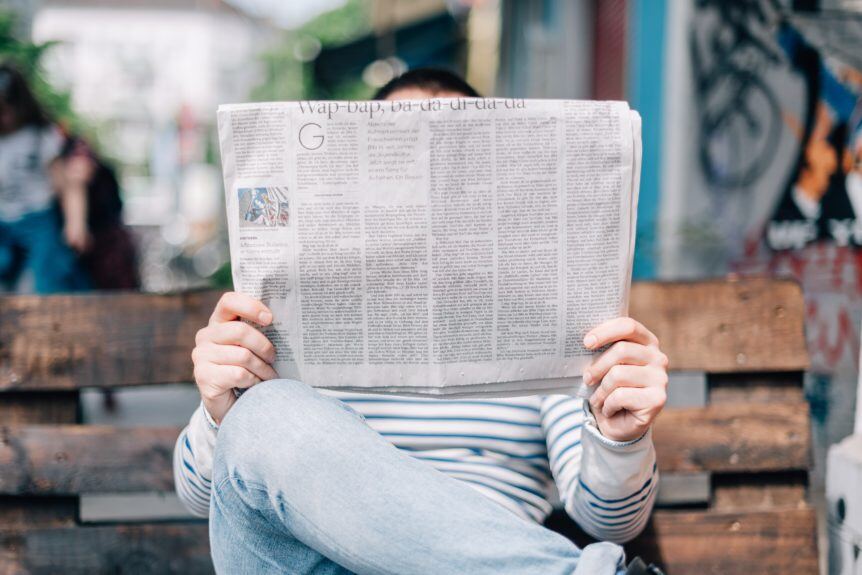 Señor sentado en un banco leyendo un periódico, cubriendo su rostro con él