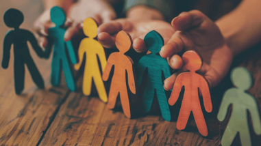 Figuras de madera de colores que representan personas protegidas por unas manos sobre una mesa de madera.