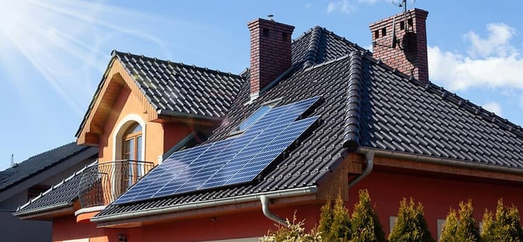 Casa con tejado de tejas oscuras y paneles solares instalados, con chimeneas de ladrillo y un cielo despejado en el fondo