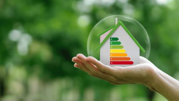 Casa con etiqueta de eficiencia energética sostenida en una mano sobre fondo natural.