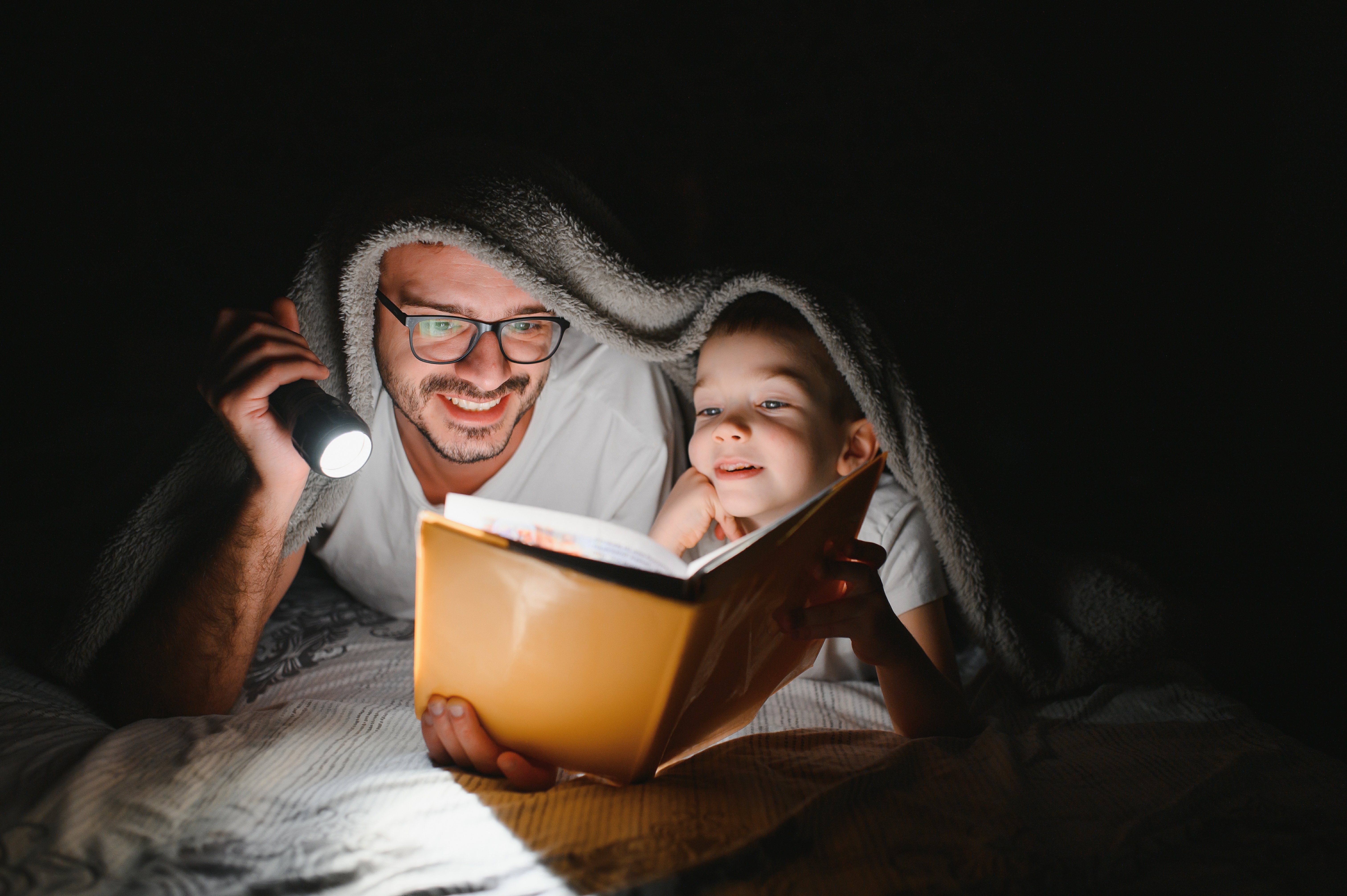 father-and-son-with-flashlight-reading-book-under-2023-11-30-21-40-47-utc