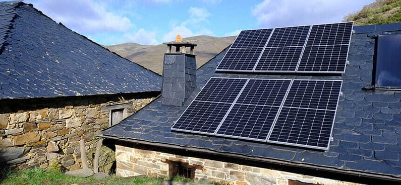 Casa de piedra con tejado de pizarra y varias placas solares instaladas, en un entorno rural con montañas al fondo