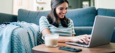Mujer sonriendo mientras trabaja con portátil en el sofá de su casa