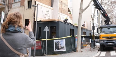 Obras de construcción en un edificio urbano con una grúa instalando elementos estructurales, mientras una persona observa y toma una fotografía del proceso.